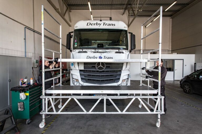 Két szakember mobil munkaállványról végzi egy Mercedes teherautó szélvédőjének szakszerű cseréjét a műhelyben.